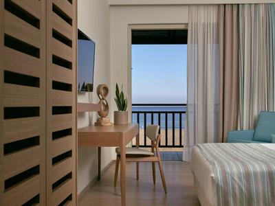 Moderní hotelový pokoj s balkonem s výhledem na moře, dřevěným stolem, židlí a útulnou postelí s pruhovaným povlečením.