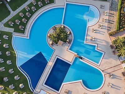 Letecký pohled na zakřivený hotelový bazén s lehátky a zelení kolem.