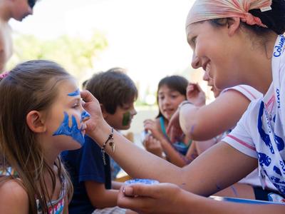 Žena s úsměvem maluje dívce obličej na cestovatelské akci.
