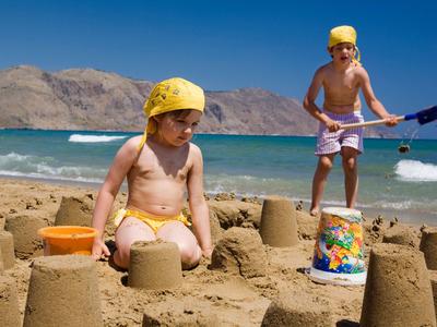 Děti staví hrady z písku na pláži za slunečného počasí s horami v pozadí.
