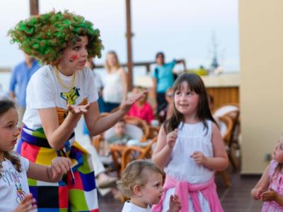 Děti slaví s klaunem na slunné hotelové terase, hravá atmosféra.