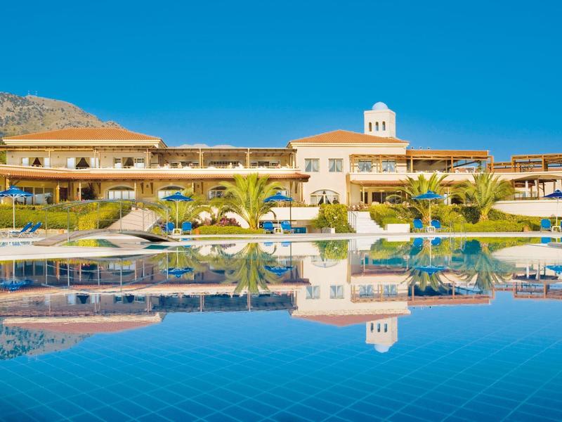 Luxusní hotel s velkým bazénem, palmami a horami pod jasně modrou oblohou.