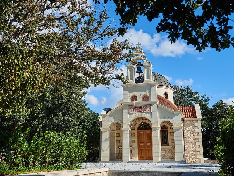 Malá bílá kaple s kupolí a zvonem, obklopená stromy pod modrou oblohou.