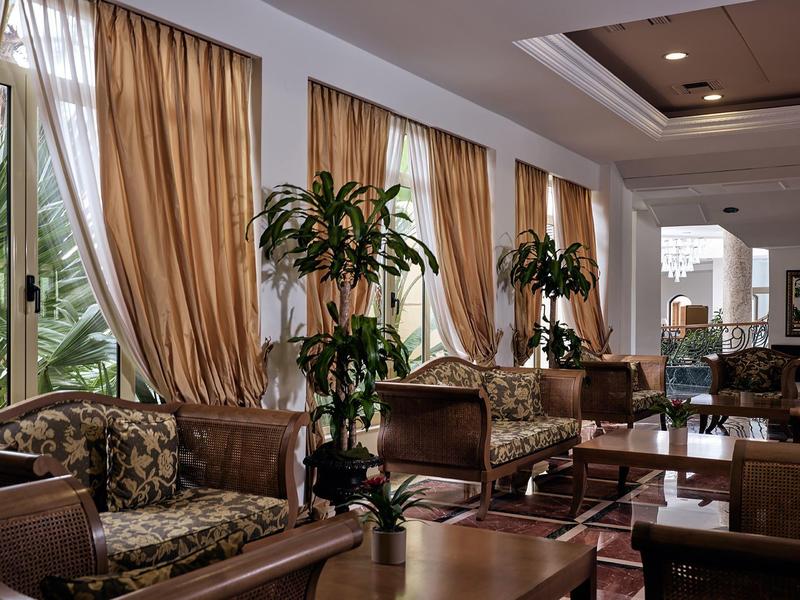 Pohodlný hotelový salonek s křesly, velkými rostlinami a zlatými závěsy.