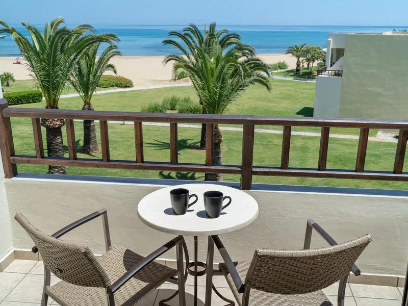 Balkon se stolem a dvěma židlemi s výhledem na palmy, zelený trávník, písečnou pláž a modré moře.