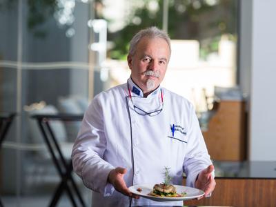 Homme âgé en tenue de chef présentant une assiette de nourriture dans un restaurant.