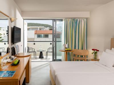 Helles Hotelzimmer mit Doppelbett, Schreibtisch und Balkon mit Blick auf andere Gebäude.