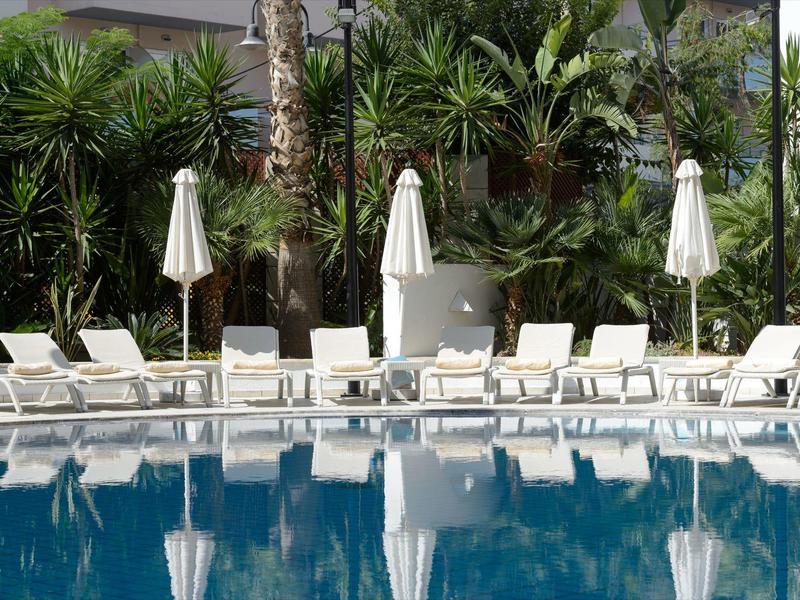Rangée de chaises longues blanches et parasols fermés au bord d'une piscine bleue claire, entourée de palmiers.