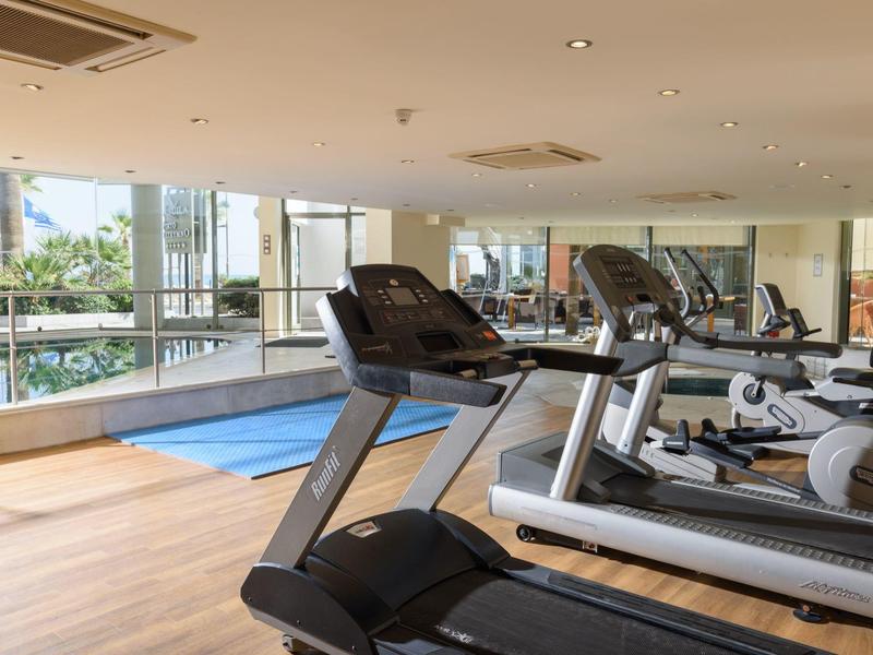 Salle de sport lumineuse avec tapis de course, grandes fenêtres et vue sur la piscine.