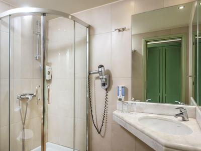 Nowoczesna łazienka hotelowa z przeszkloną kabiną prysznicową, umywalką, lustrem, suszarką i zielonymi drzwiami w odbiciu.