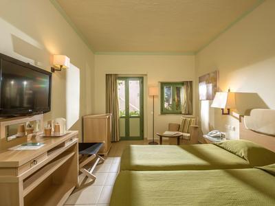 Pokój hotelowy z dwoma zielonymi łóżkami, biurkiem, TV i miejscem do siedzenia przy drzwiach na balkon.