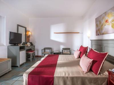 Jasny pokój hotelowy z dwoma łóżkami, czerwonymi akcentami, telewizorem i biurkiem przy oknie.