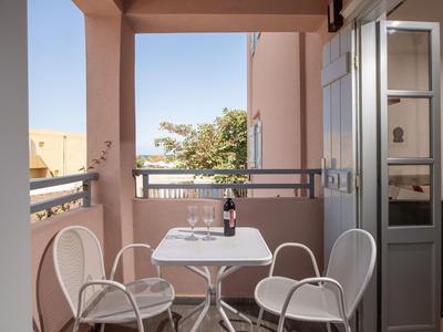 Balkon se čtyřmi bílými židlemi, stolem, lahví vína a dvěma sklenicemi na pozadí modré oblohy.