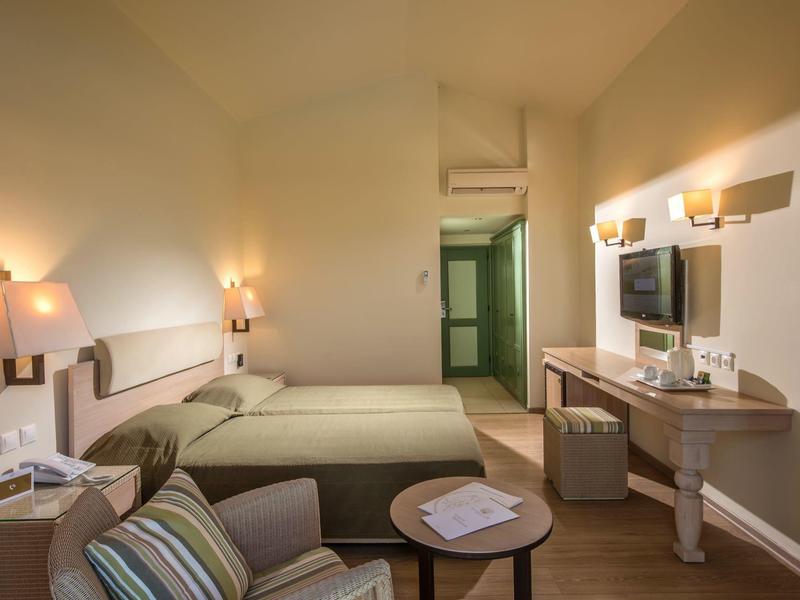 Przytulny pokój hotelowy z dwoma łóżkami, biurkiem z telewizorem, fotelem i miękkim oświetleniem.