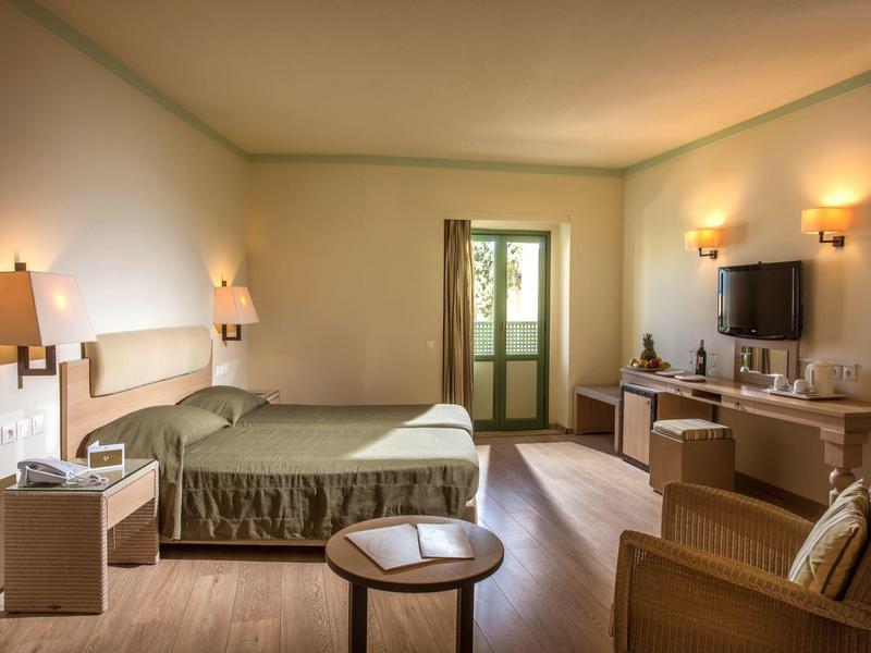 Przestronny pokój hotelowy z podwójnym łóżkiem, fotelem, telewizorem i drzwiami na balkon.