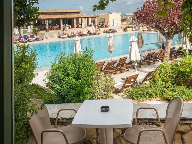 Strefa zewnętrzna z stołem, krzesłami i widokiem na basen oraz leżaki pod parasolami.