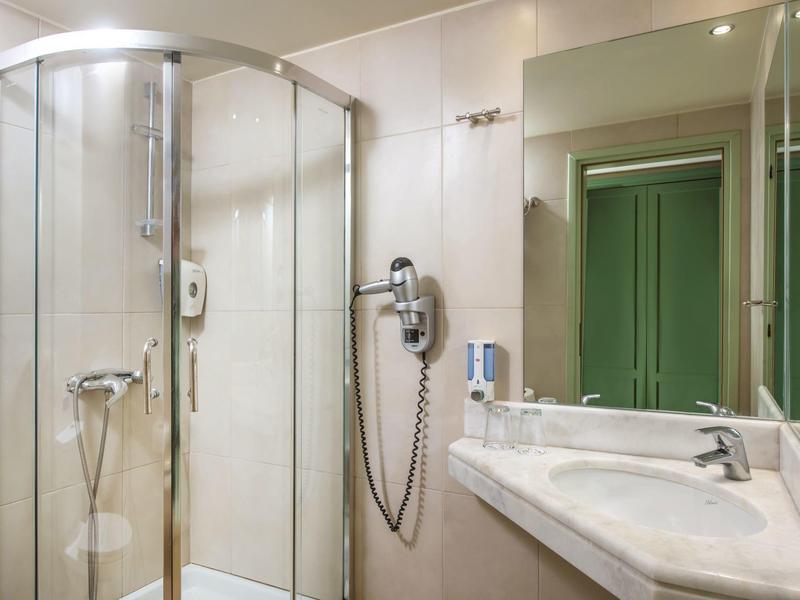 Nowoczesna łazienka hotelowa z przeszkloną kabiną prysznicową, umywalką, lustrem, suszarką i zielonymi drzwiami w odbiciu.