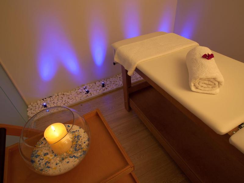 Przytulny pokój spa z łóżkiem do masażu, zrolowanym ręcznikiem, kwiatem, świecą i niebieskim oświetleniem ściennym.