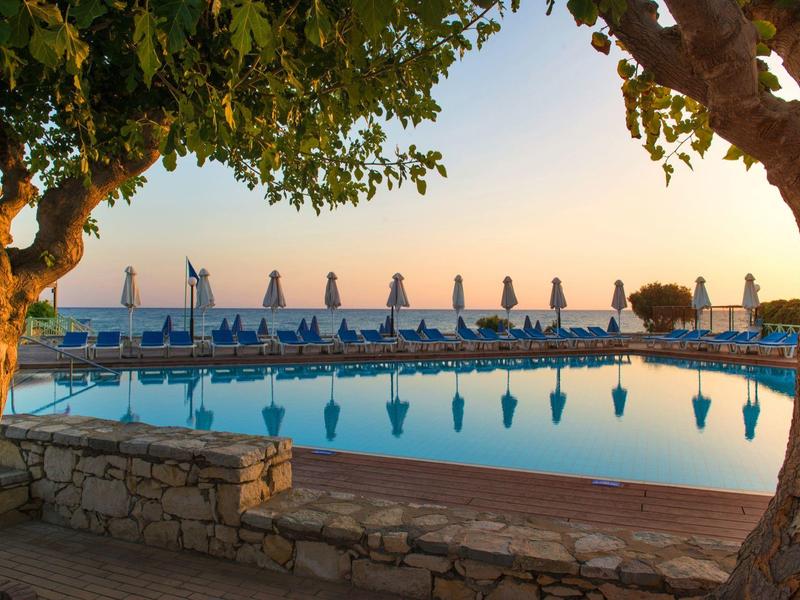 Zachód słońca nad spokojnym basenem z parasolami i drzewami na pierwszym planie.