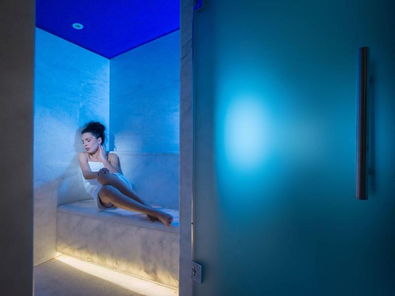Kobieta relaksuje się w nowoczesnej łaźni parowej z niebieskim oświetleniem i szklanymi drzwiami.