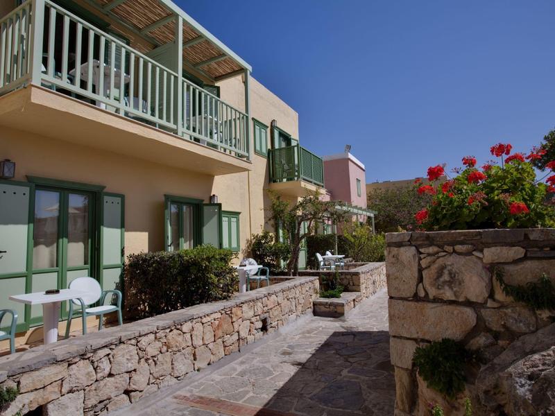 Słoneczna elewacja hotelu z balkonami, białymi krzesłami, kamiennymi murami i czerwonymi kwiatami.