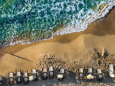 Toma aérea de playa con filas de tumbonas y mar verdeazul con olas blancas.