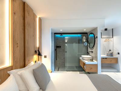 Habitación de hotel moderna con cama doble, detalles de madera y ducha de vidrio al fondo.