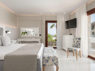 Habitación de hotel luminosa con cama doble grande, banco, televisor y balcón con vista a palmeras.