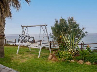 Jardín con banco columpio junto al mar, palmeras y césped bien cuidado bajo cielo despejado.