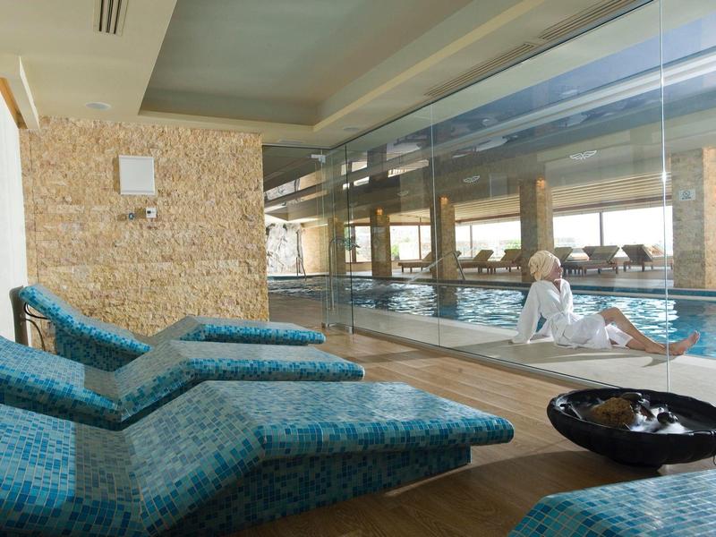 Área de piscina interior con tumbonas y una gran ventana que deja entrar la luz diurna.