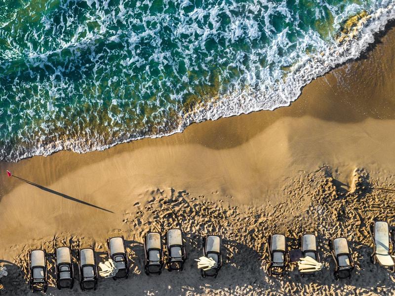 Toma aérea de playa con filas de tumbonas y mar verdeazul con olas blancas.
