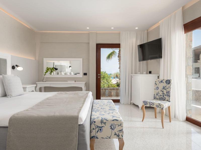 Habitación de hotel luminosa con cama doble grande, banco, televisor y balcón con vista a palmeras.