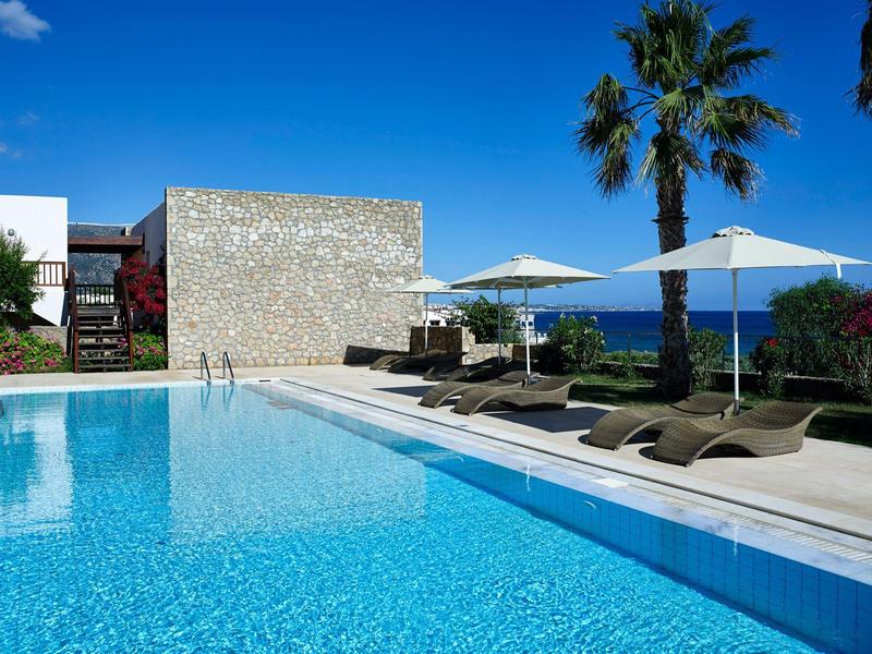 Piscina exterior moderna con tumbonas y palmeras bajo cielo azul junto al mar.