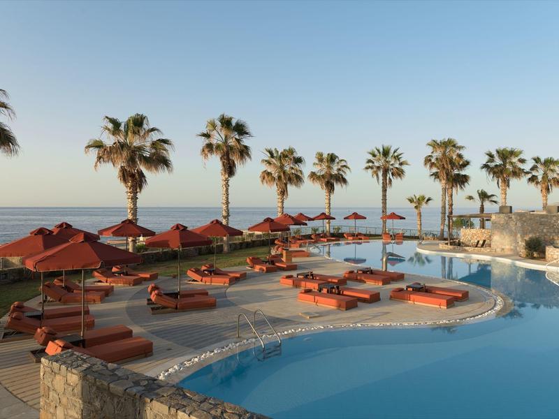 Vista de una zona de piscina con tumbonas, sombrillas y palmeras junto al mar bajo un cielo despejado.