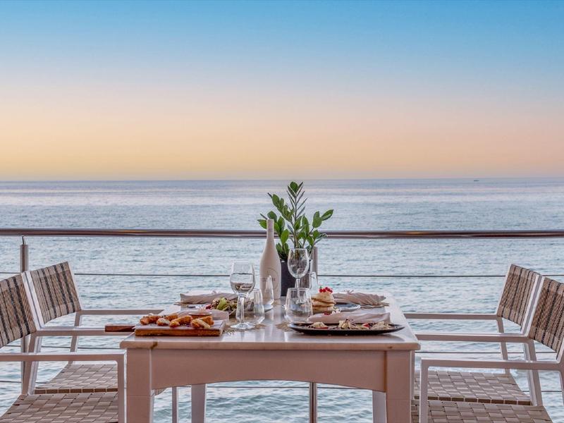 Mesa de comedor en balcón con vista al mar y atardecer, con platos y vasos servidos.