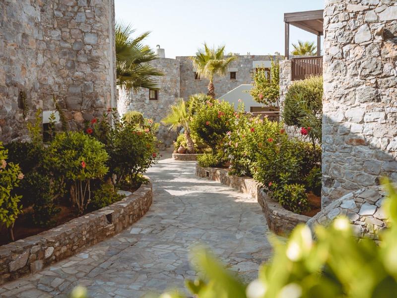 Un camino pavimentado entre muros bajos de piedra atraviesa un jardín verde con palmeras y arbustos.