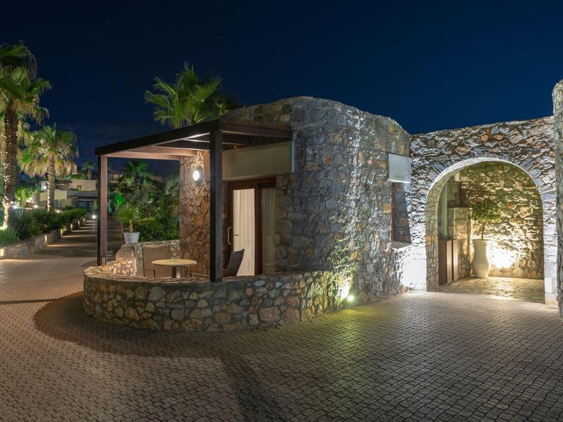 Entrada y terraza de piedra iluminadas en un hotel por la noche con palmeras al fondo.