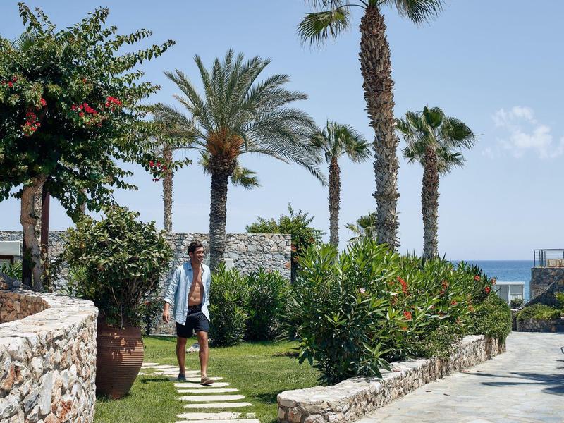 Hombre caminando por un jardín tropical con palmeras y vista al mar bajo cielo azul claro