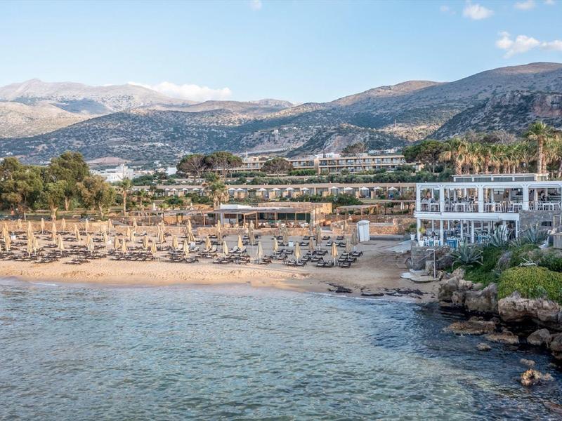 Hotel de playa con tumbonas, palmeras y montañas al fondo bajo un cielo azul.