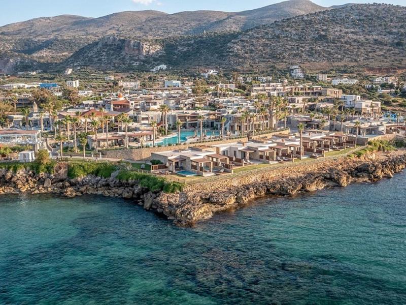 Complejo hotelero en la costa con piscina, rodeado de montañas y agua azul clara.