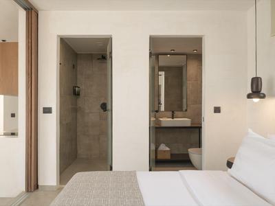 Modern hotel room with white bed, beige rug, concrete shower and a sink with mirror.