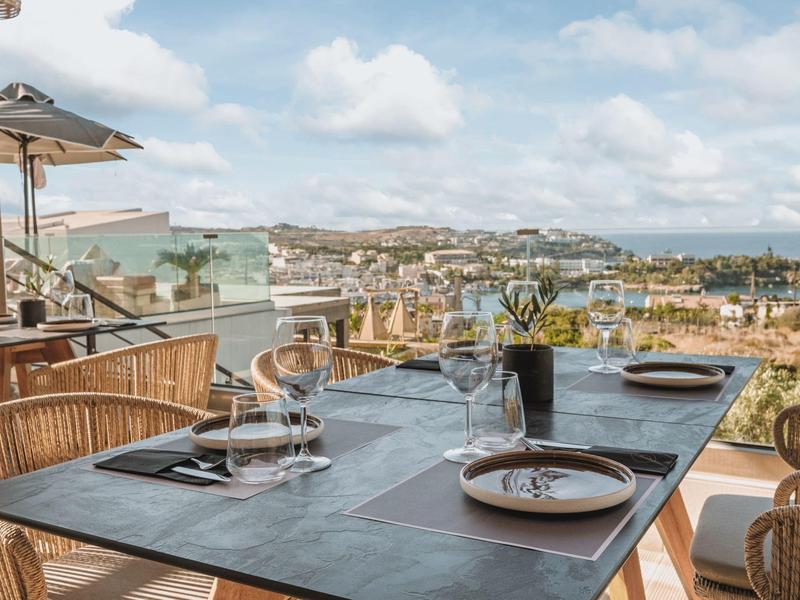 Table set for four with wine glasses and plates on a terrace overlooking a coastal town.