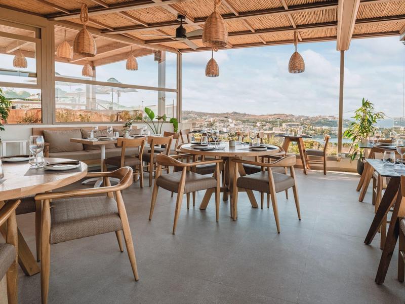 Bright hotel restaurant with wooden furniture and panoramic city views through large windows.