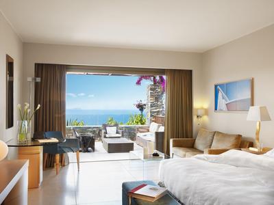 Helles Hotelzimmer mit Bett und Sitzbereich, Blick auf eine Terrasse und das Meer im Hintergrund.