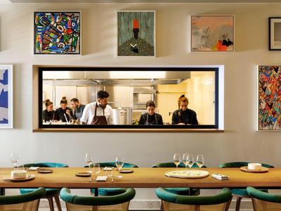 Pohled na moderní restauraci s prostřenými stoly a kuchyňským personálem přes obdélníkové okno.