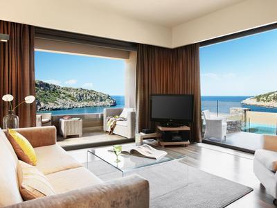 Světý obývací pokoj s velkými okny s výhledem na moře a terasu pod modrou oblohou.