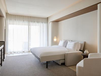 Helles Hotelzimmer mit Doppelbett, weißen Möbeln und großen Fenstern mit Vorhängen.