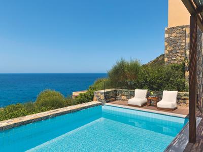 Pool mit Liegestühlen neben einem Gebäude und Blick auf das Meer bei klarem Himmel.