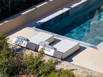 Zwei weiße Liegen neben einem klaren Pool mit Wasser, umgeben von Pflanzen und Steinboden.