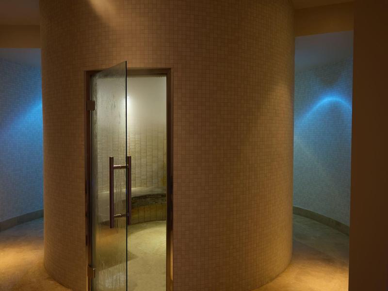 Moderní hotelová chodba se zakřivenou strukturální stěnou a skleněnými dveřmi za tlumeného osvětlení.